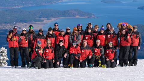 A large group of Schweitzer Ski Patrol standing at the summit with a stunning view of Lake Pend Oreille in the distance. 