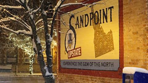 Sign painted on the side of a building downtown saying "Sandpoint: Experience the Gem of the North" with snow and holiday lights